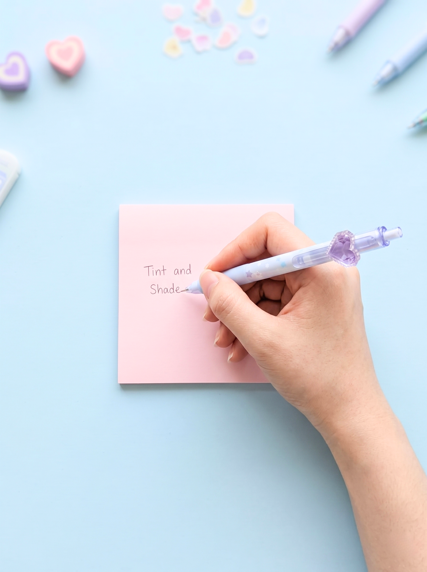 Hand holding a pen over a pink notepad with colorful heart-shaped objects on a light blue background