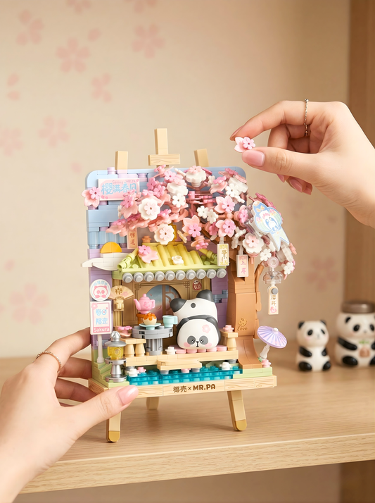 Hand holding a small, detailed model of a house with cherry blossoms on a wooden stand.