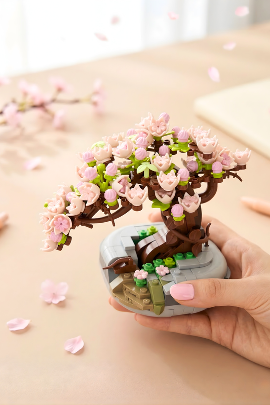 Hand holding a small model of a cherry blossom tree with pink flowers on a light surface.
