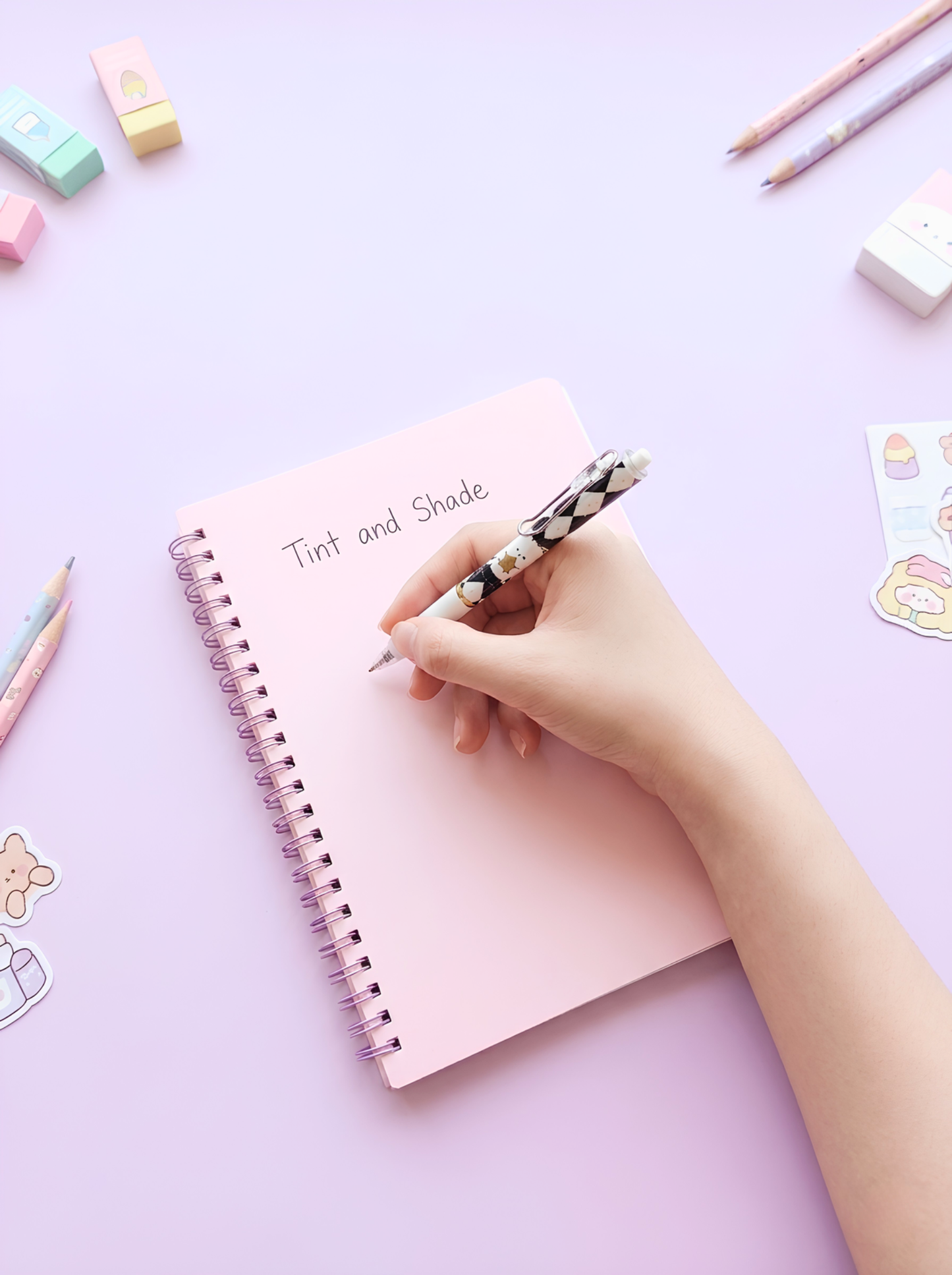Hand holding a pen over a pink notebook on a light purple background with stationery items.