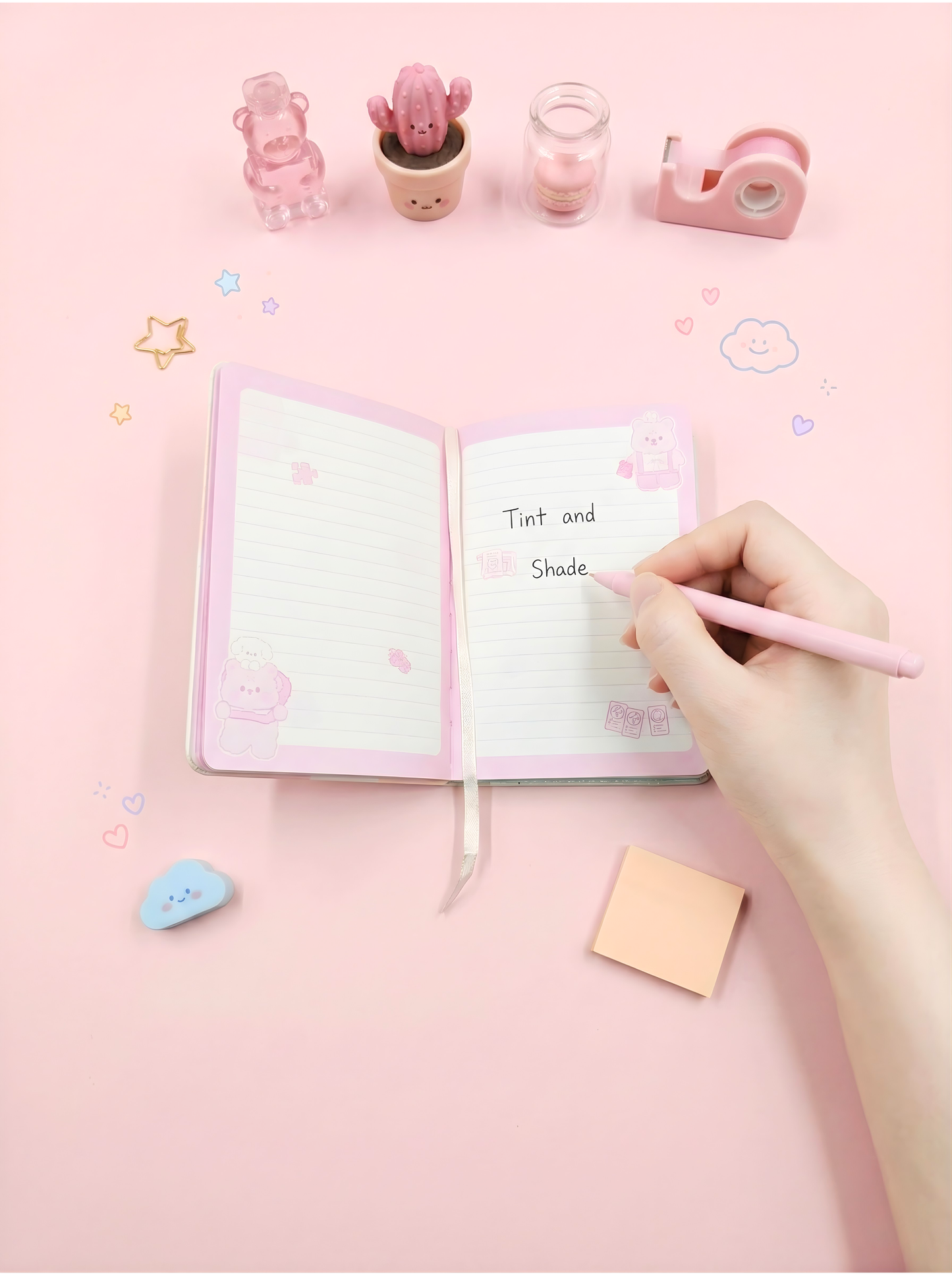 Open notebook with a hand holding a pink pen on a pink background with stationery items.