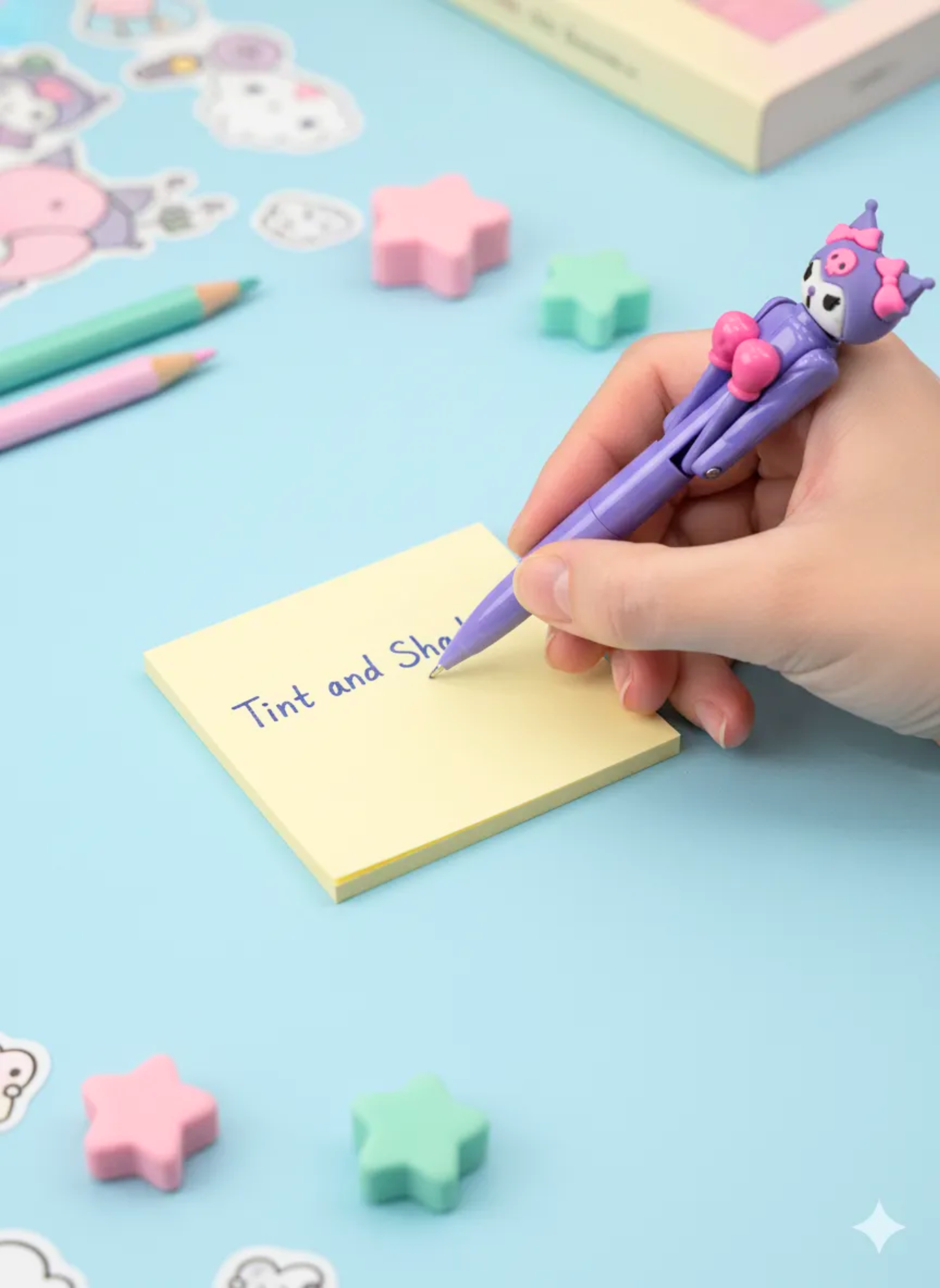 Hand holding a purple cartoon-style pen with a pink character on a blue background with colorful stationery items.