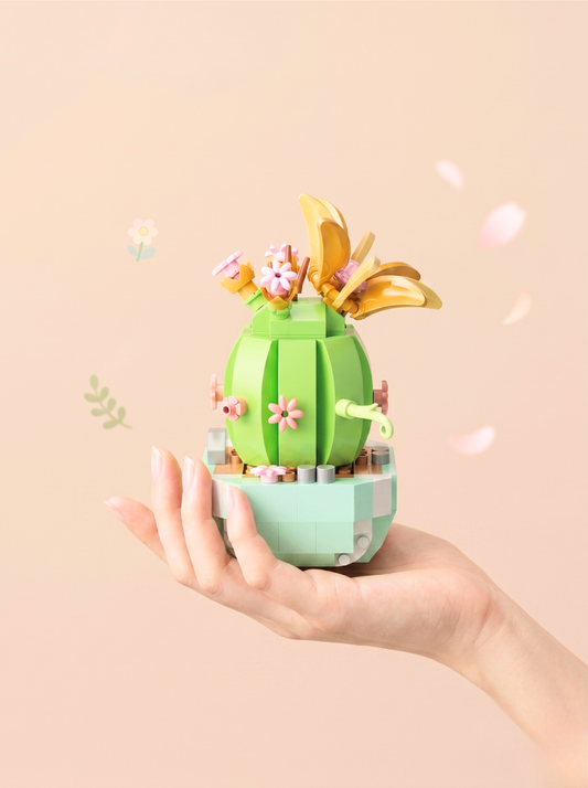 Hand holding a small green cactus-like toy with flowers and gold leaves on a beige background