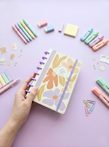 Floral notebook held by a hand with stationery items on a light purple background