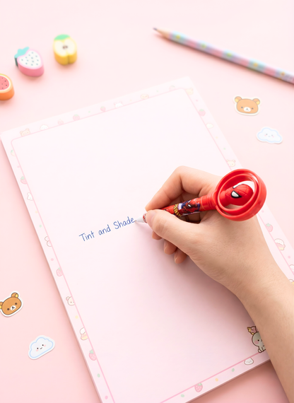 Hand holding a red pen with a spider design, writing on a pink notebook with stickers around.