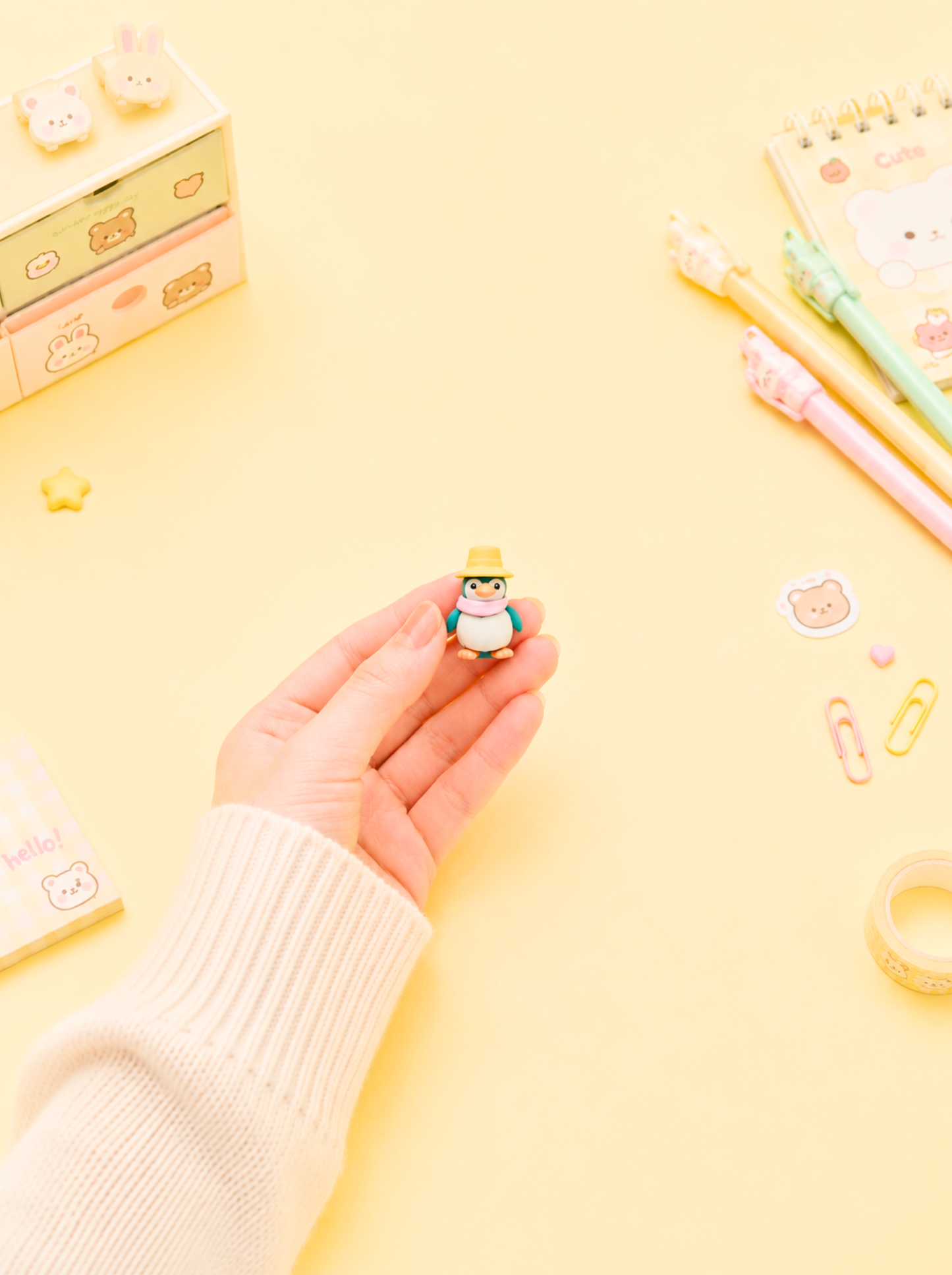 Hand holding a small figurine on a yellow background with stationery items