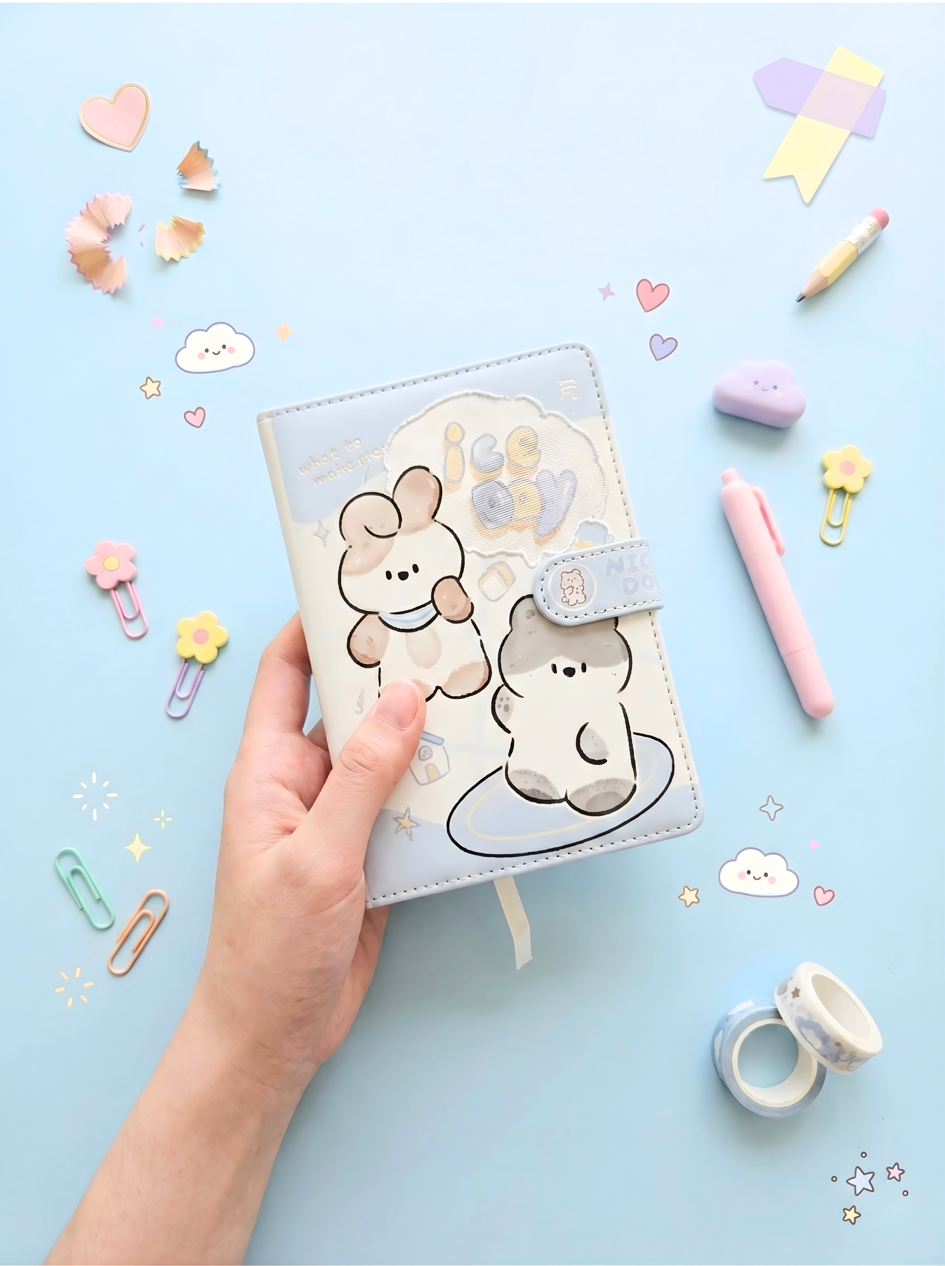 Hand holding a small notebook with bear illustrations on a light blue background with stationery items.