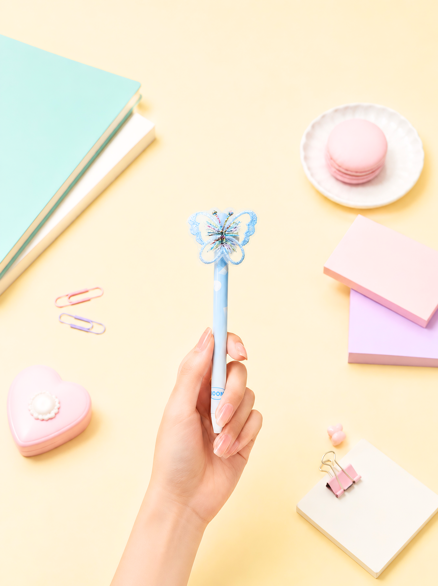 Hand holding a blue butterfly pen on a yellow surface with stationery items.