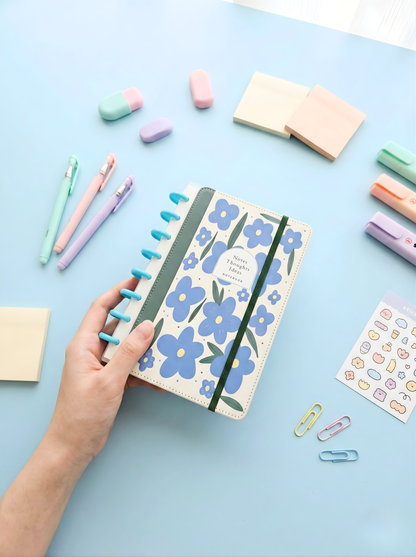 Floral notebook held by a hand on a light blue background with stationery items.
