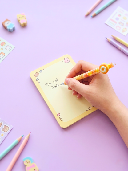 Hand writing on a yellow notepad with colorful stationery items on a purple background
