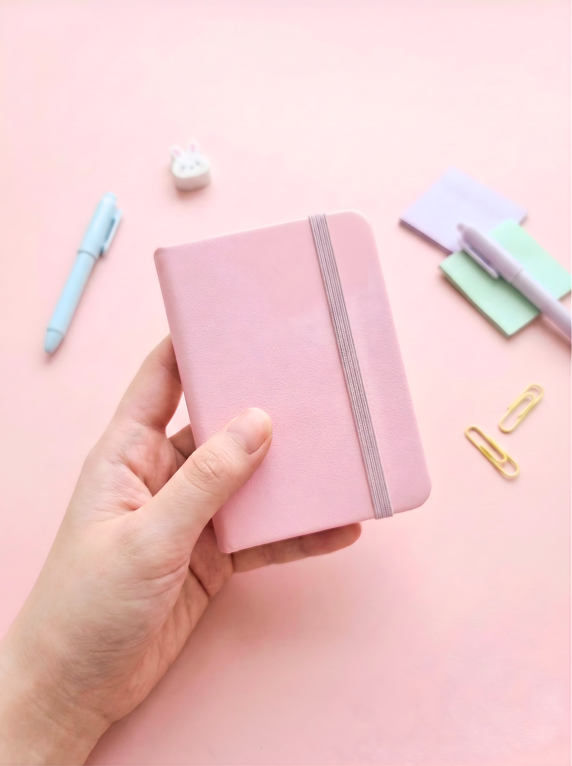 Hand holding a pink notebook with stationery items on a pink background
