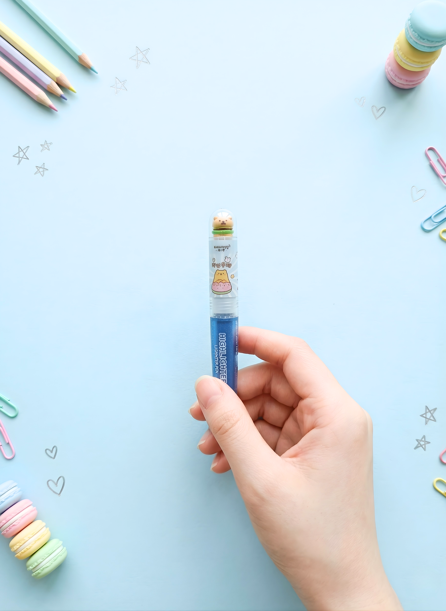 Hand holding a blue highlighter with cartoon character on a light blue background with colorful pencils and paper clips.