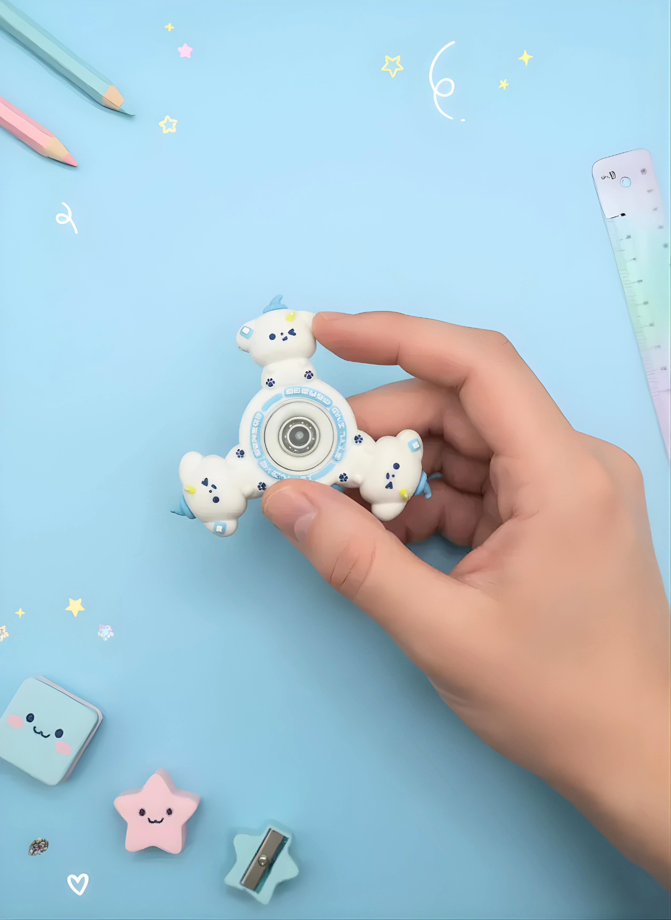 Hand holding a white fidget spinner with bear designs on a light blue background with stationery items.