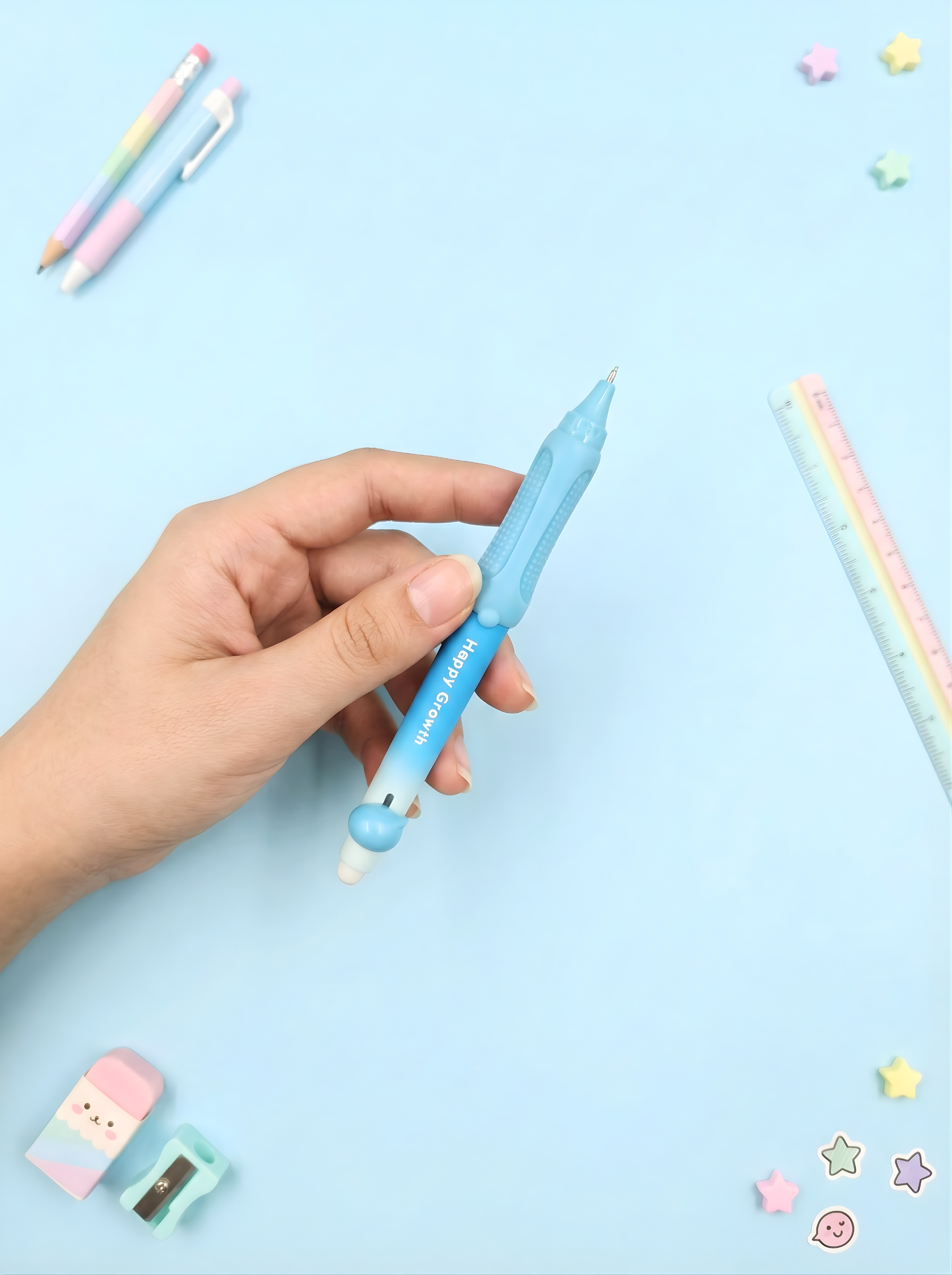 Hand holding a blue pen with a cartoon character on a light blue background with stationery items.