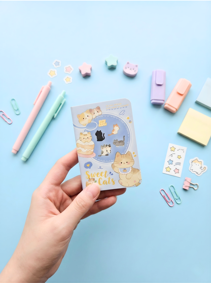 Hand holding a small notebook with 'Sweet Cats' design on a pink background with stationery items.