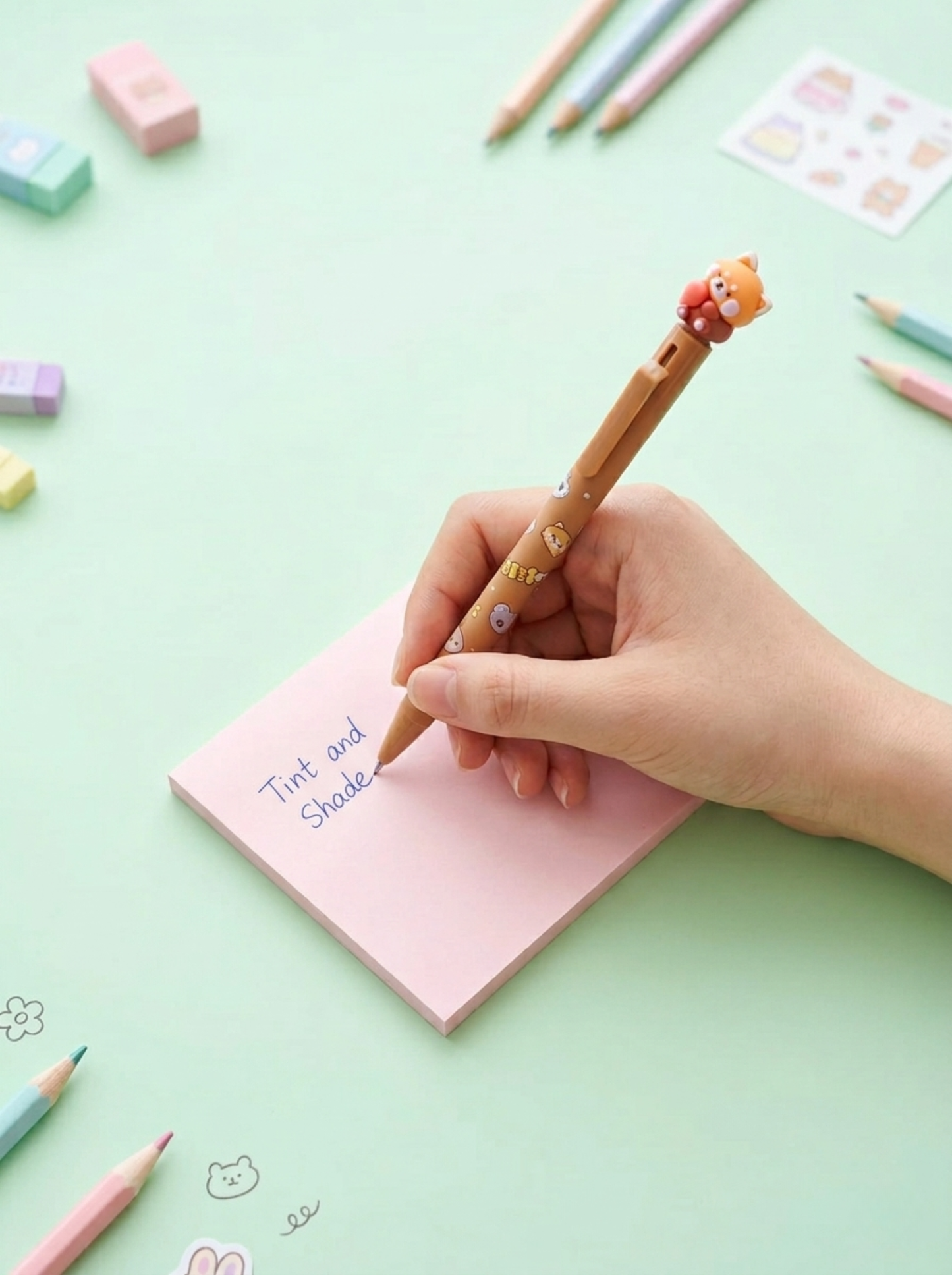 Hand holding a decorative pen over a pink notepad with stationery items on a light green background