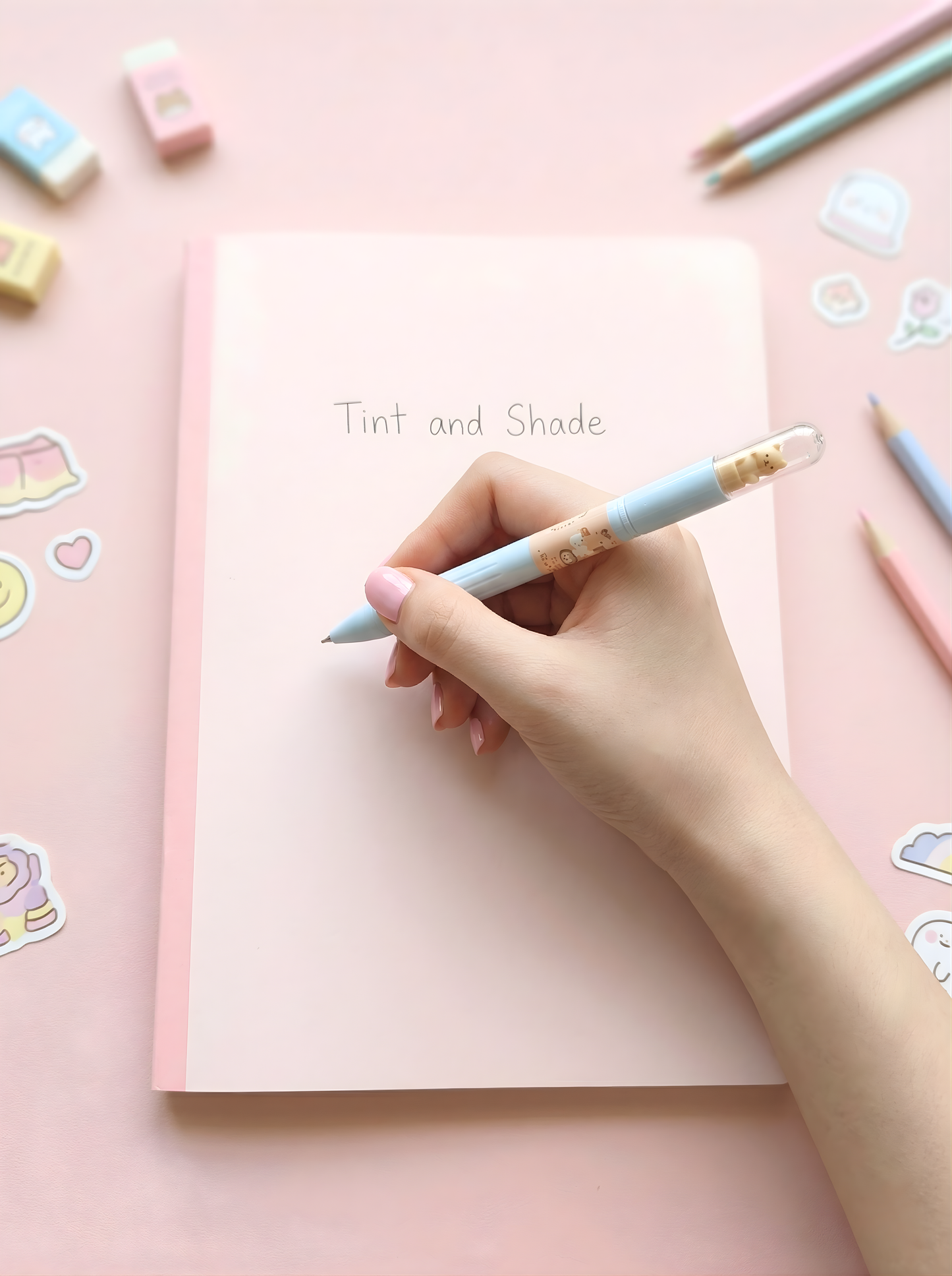 Hand holding a light blue pencil over a pink notebook with stickers on a pink background