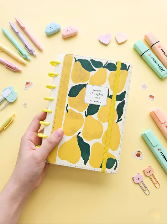 Notebook with pear design held by a hand on a yellow background with stationery items.