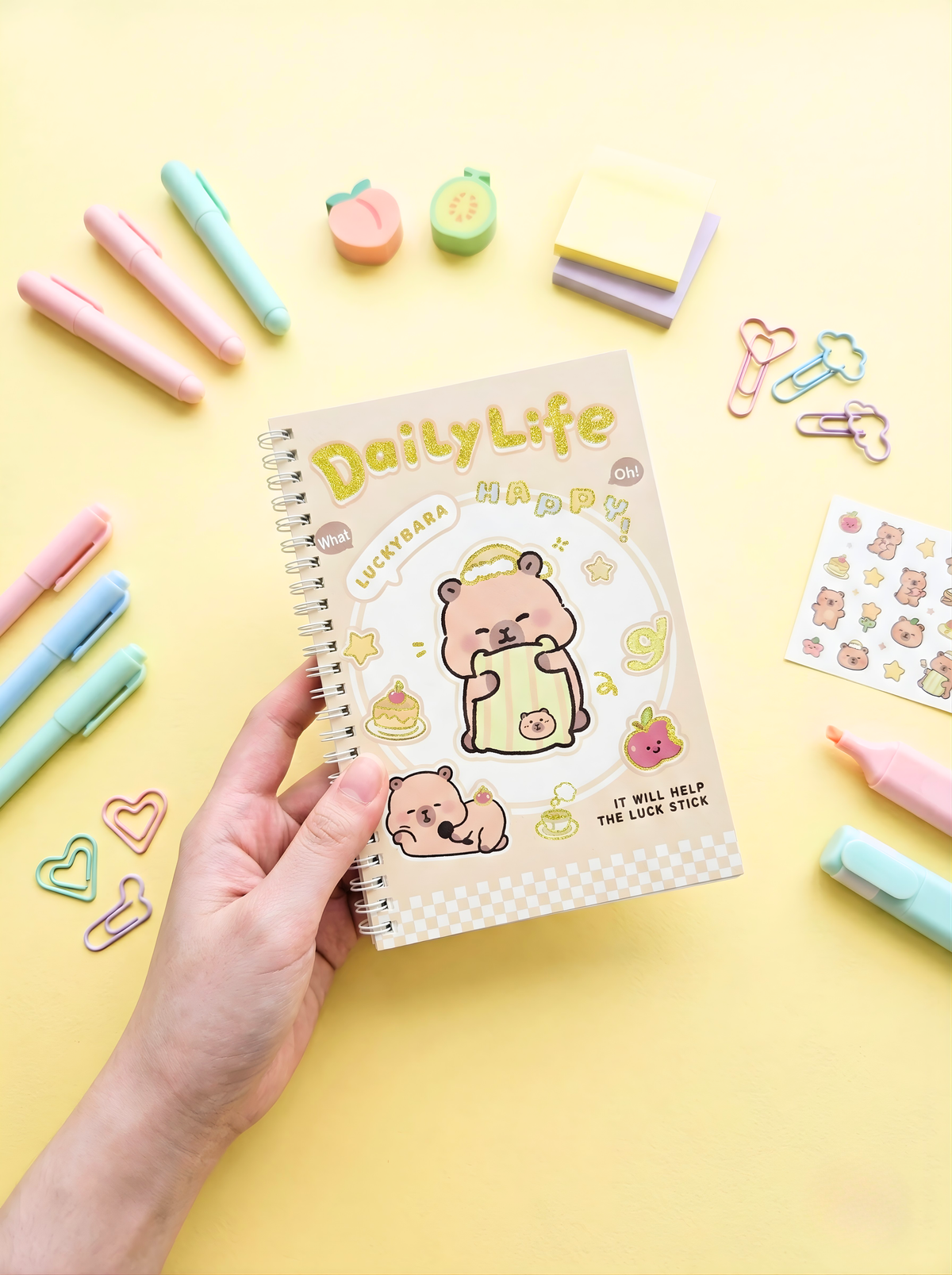 Notebook titled 'Daily Life' with a bear illustration, held by a hand on a yellow background with stationery items.