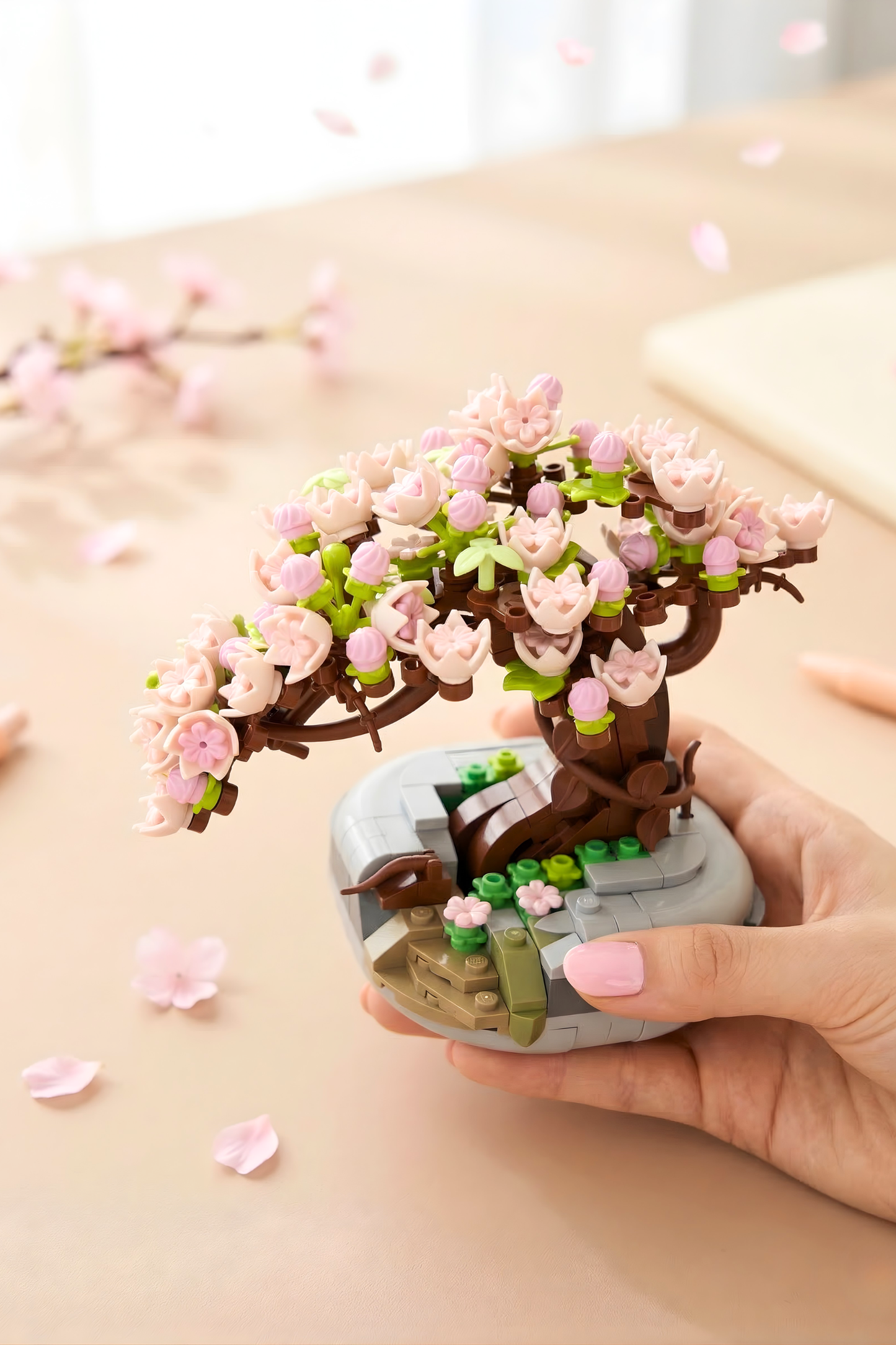 Hand holding a small model of a cherry blossom tree with pink flowers on a light surface.