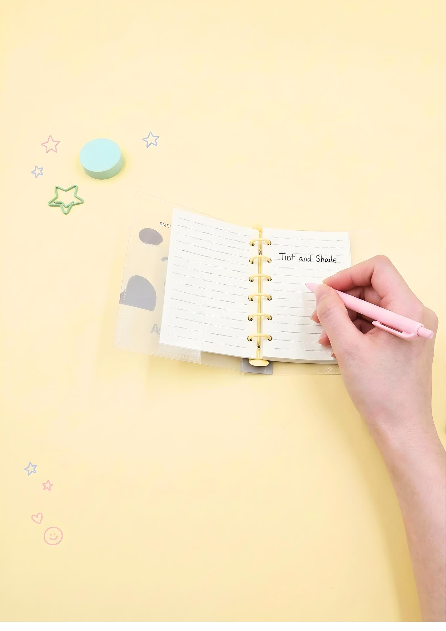 Hand holding a pink pen over an open notebook on a yellow background with decorative stickers.