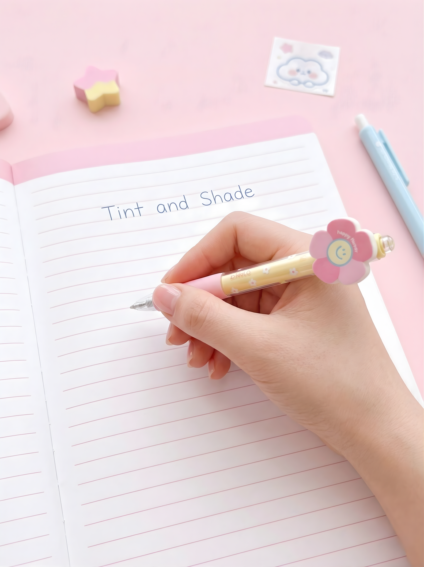 Hand holding a pen with a decorative flower top, writing in a notebook on a pink surface.