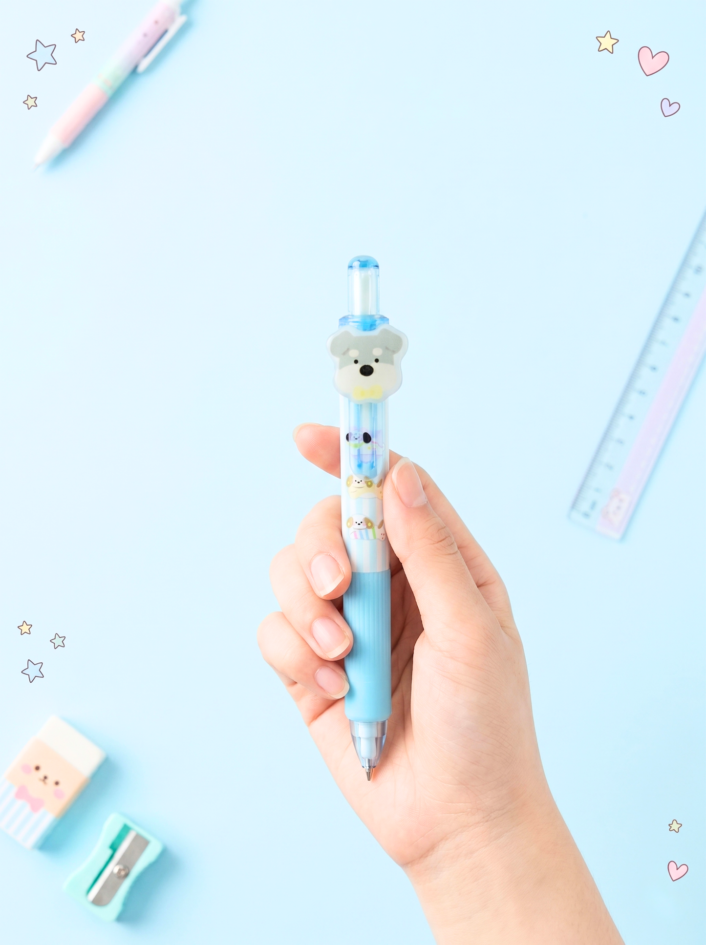 Hand holding a blue cartoon-themed pen on a light blue background with stationery items.