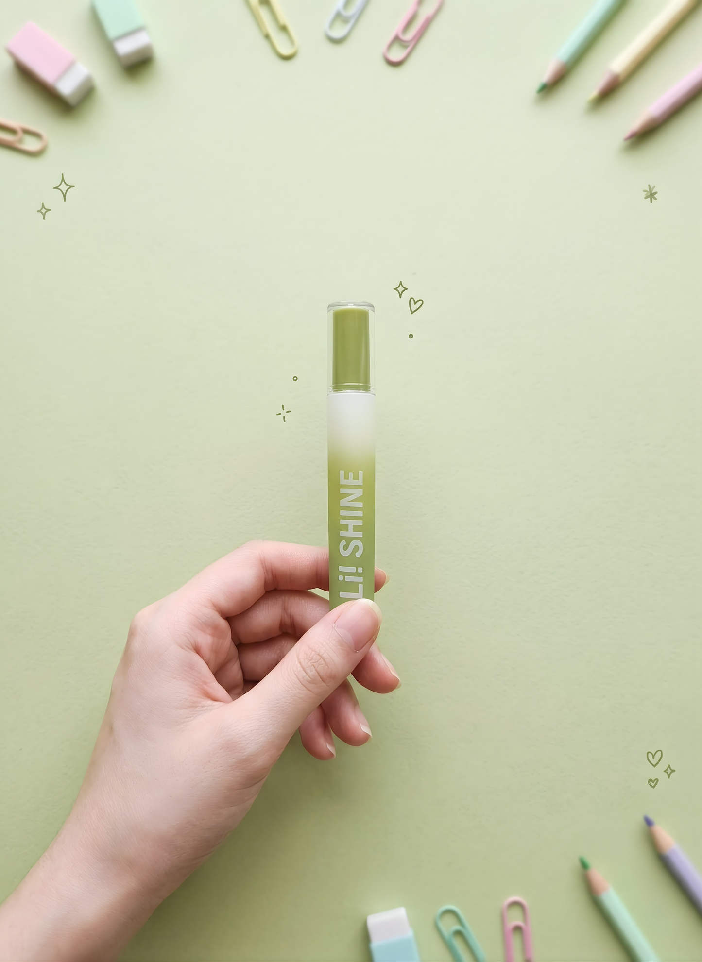Hand holding a green and white pen with 'SHINE' branding on a light green background with stationery items.