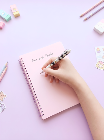 Hand holding a pen over a pink notebook on a light purple background with stationery items.