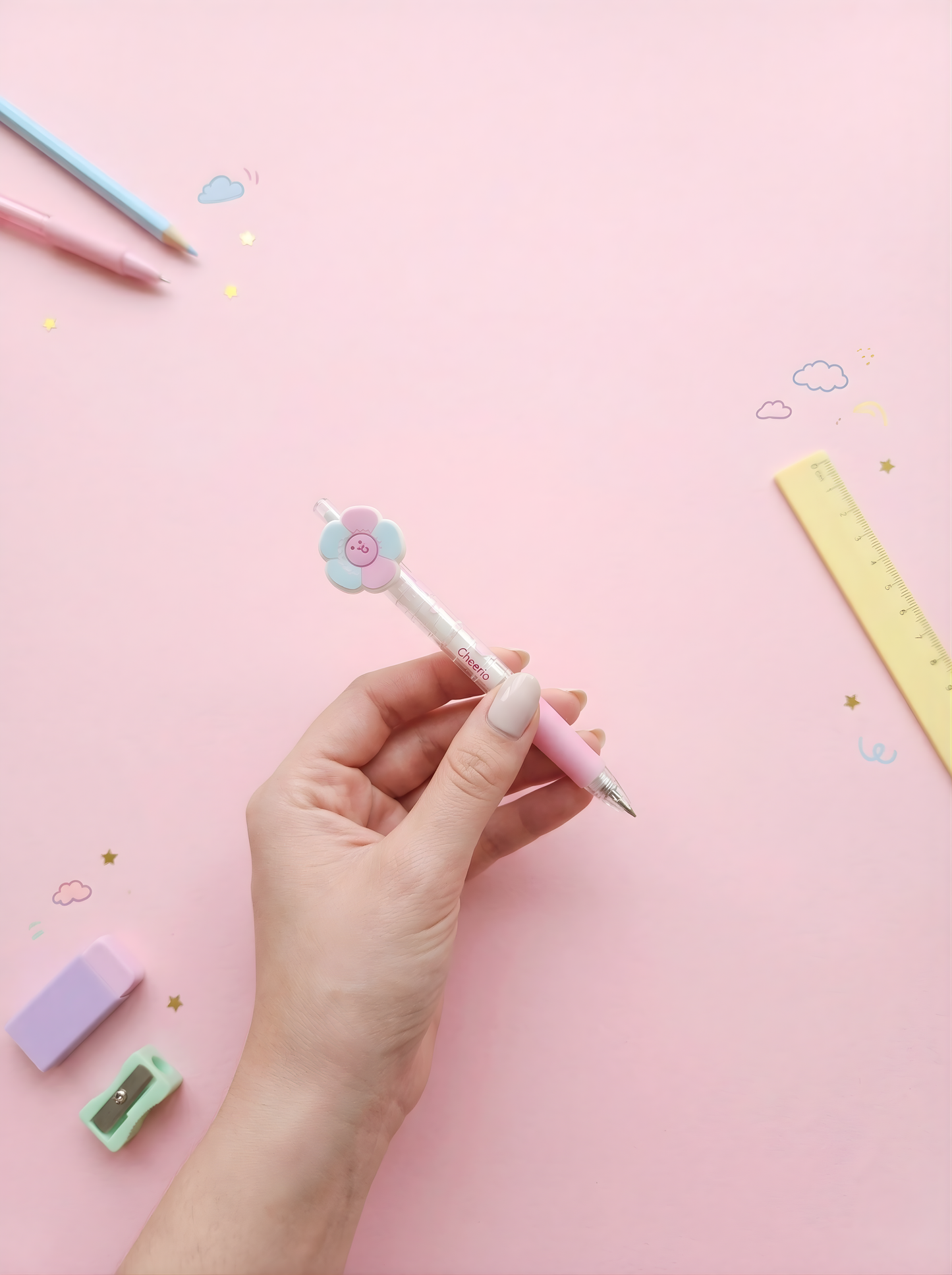 Hand holding a pink pen with a flower design on a pink background with stationery items.