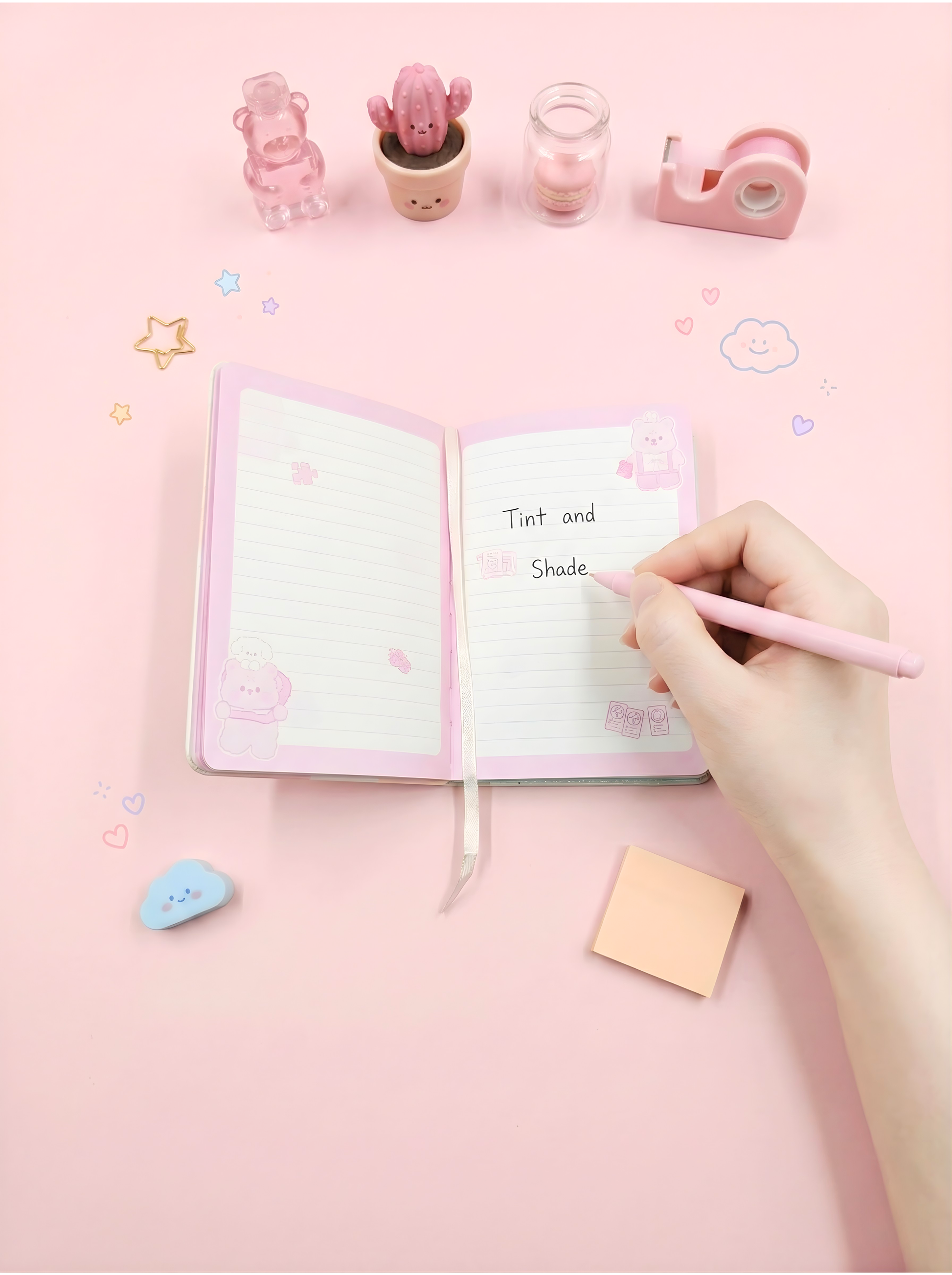 Open notebook with a hand holding a pink pen on a pink background with stationery items.