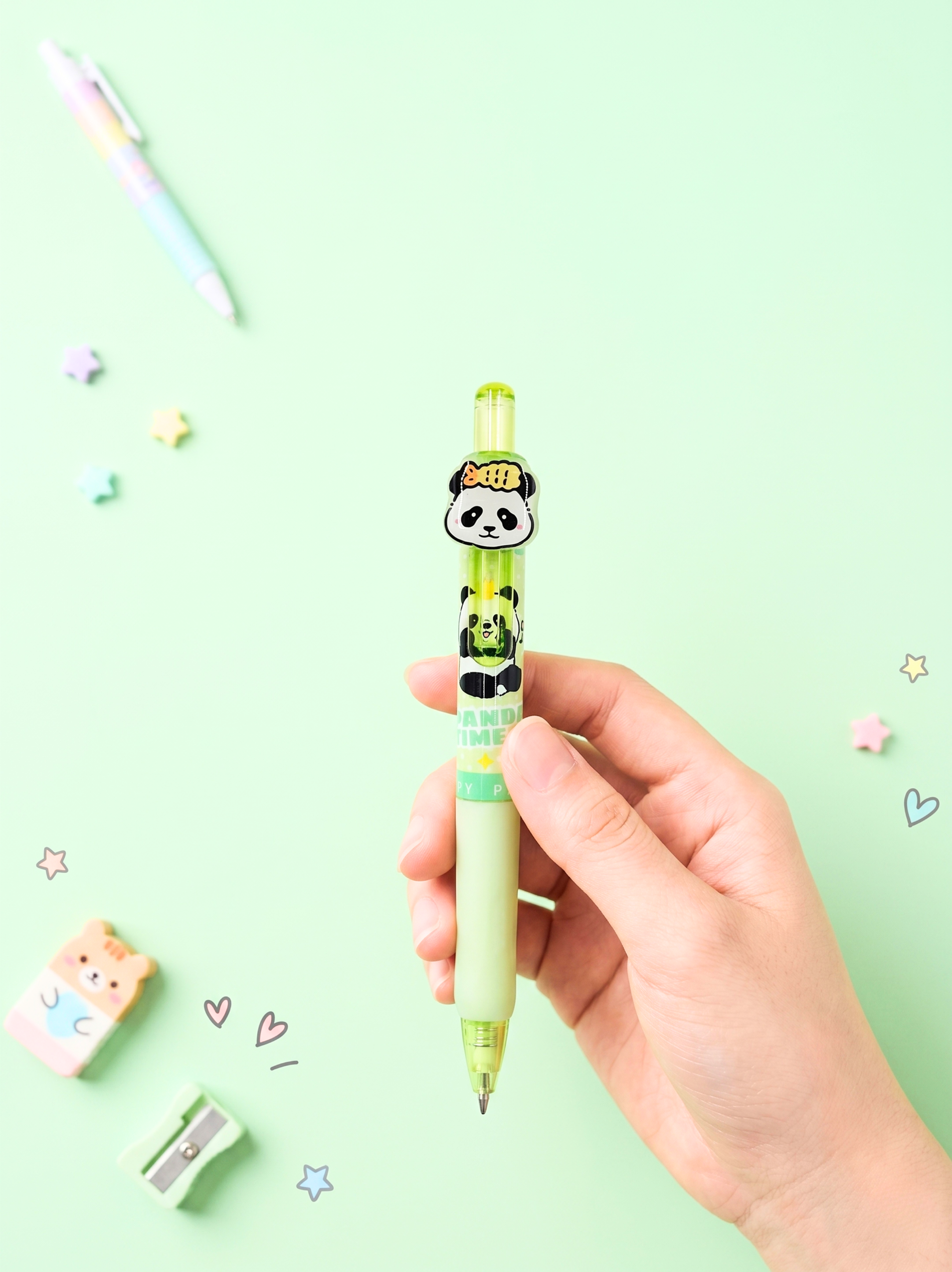 Hand holding a panda-themed mechanical pencil against a light green background with stationery items.