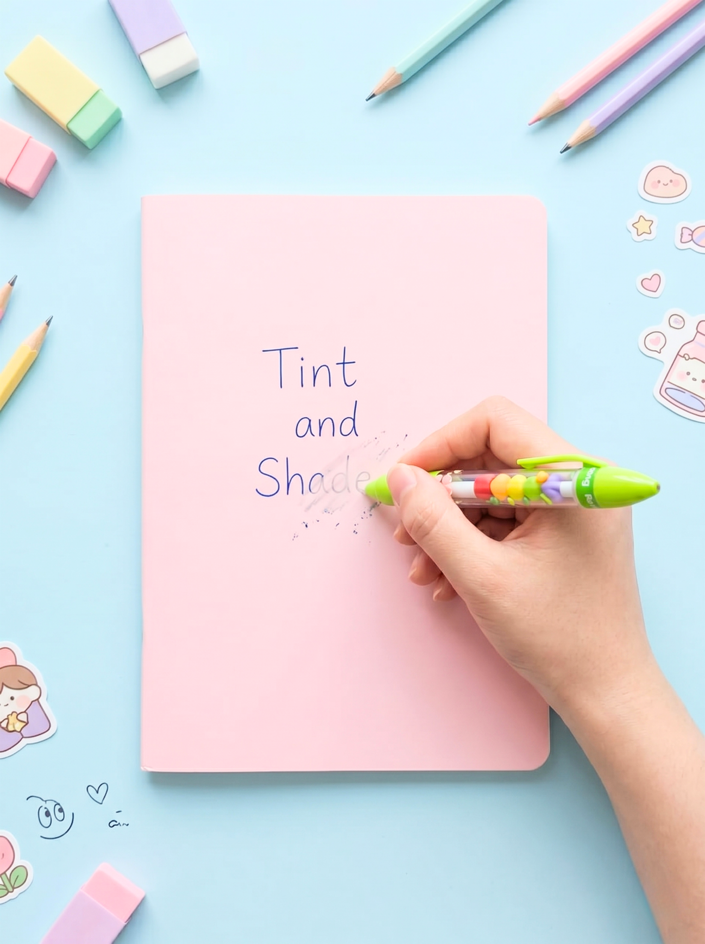 Hand writing 'Tint and She' on a pink notebook with colorful pens and markers around.