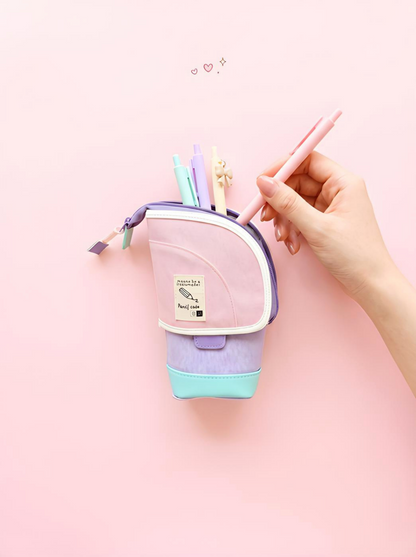 Hand holding a pink pen over a small purple pouch with pens on a pink background