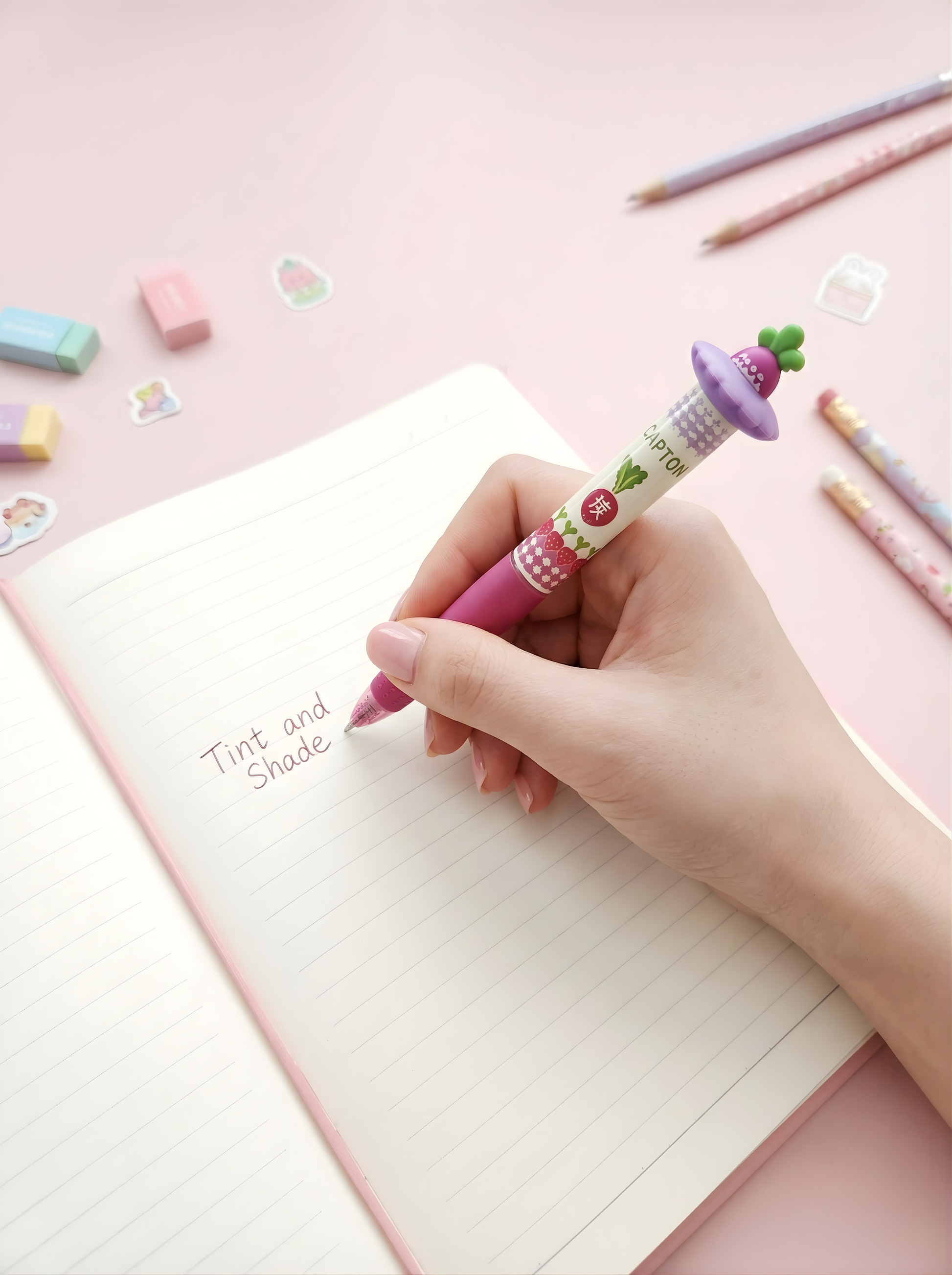 Hand holding a pink pen with a cartoon character on a pink notebook with stationery items in the background.