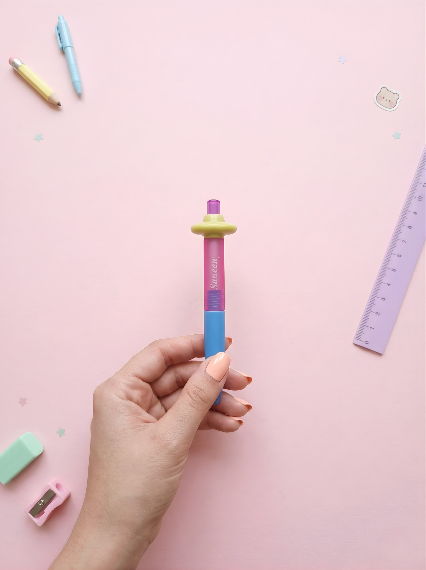 Colorful pen held by a hand on a pink background with stationery items.