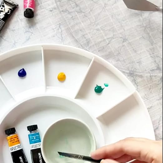 Paint palette with colorful paint samples and a hand holding a brush on a light gray surface.