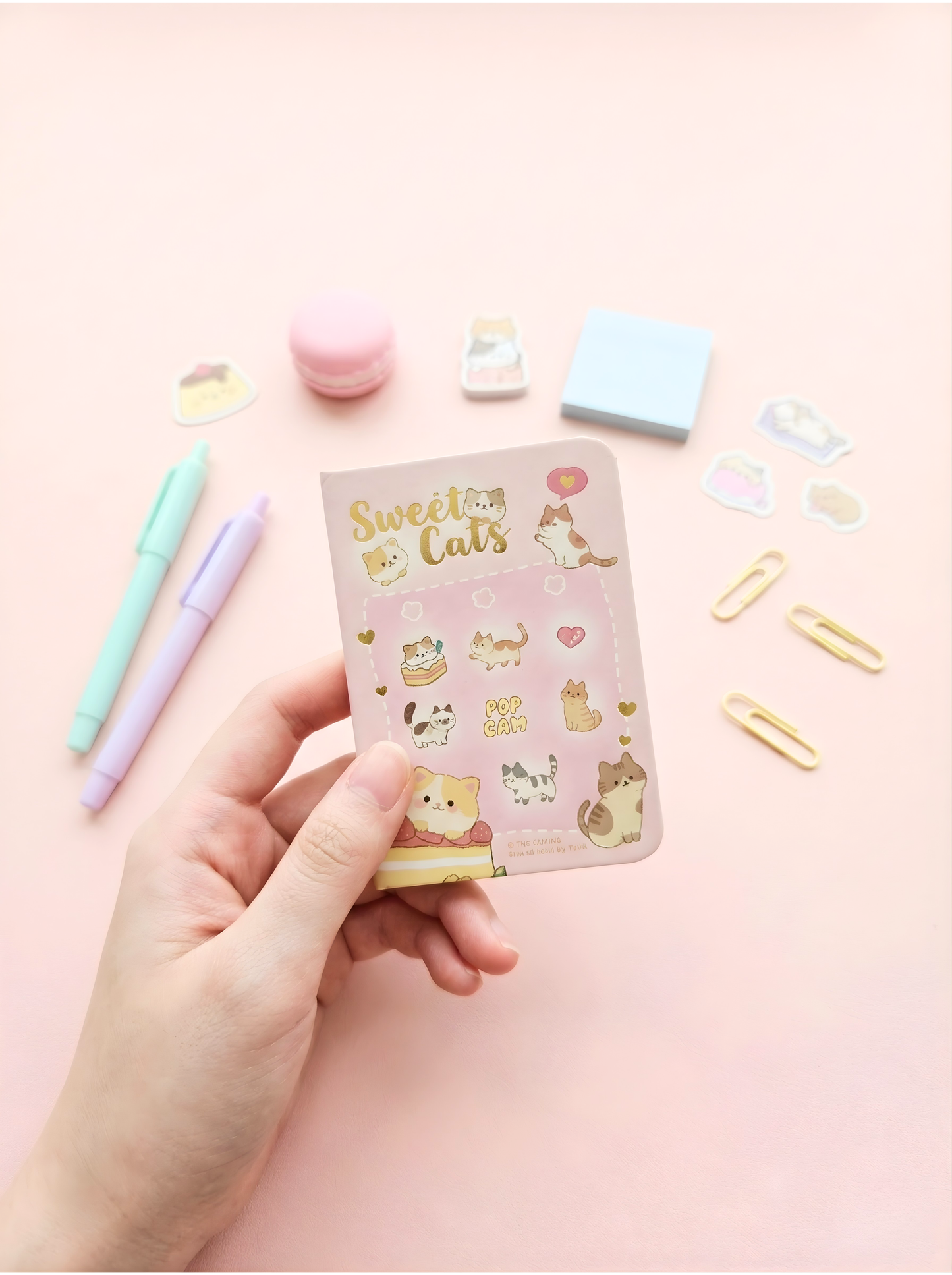 Hand holding a small notebook with 'Sweet Cats' design on a pink background with stationery items.