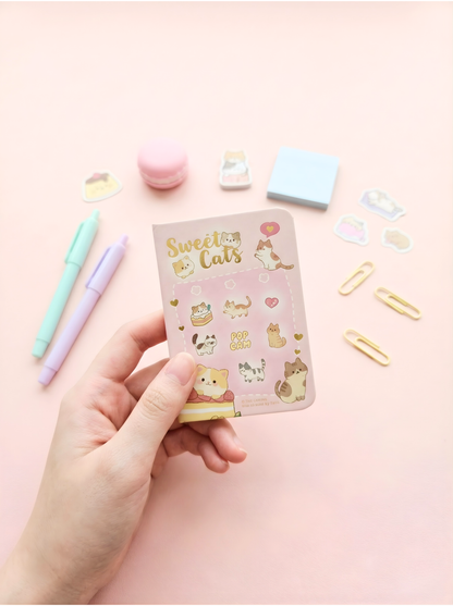 Hand holding a small notebook with 'Sweet Cats' design on a pink background with stationery items.