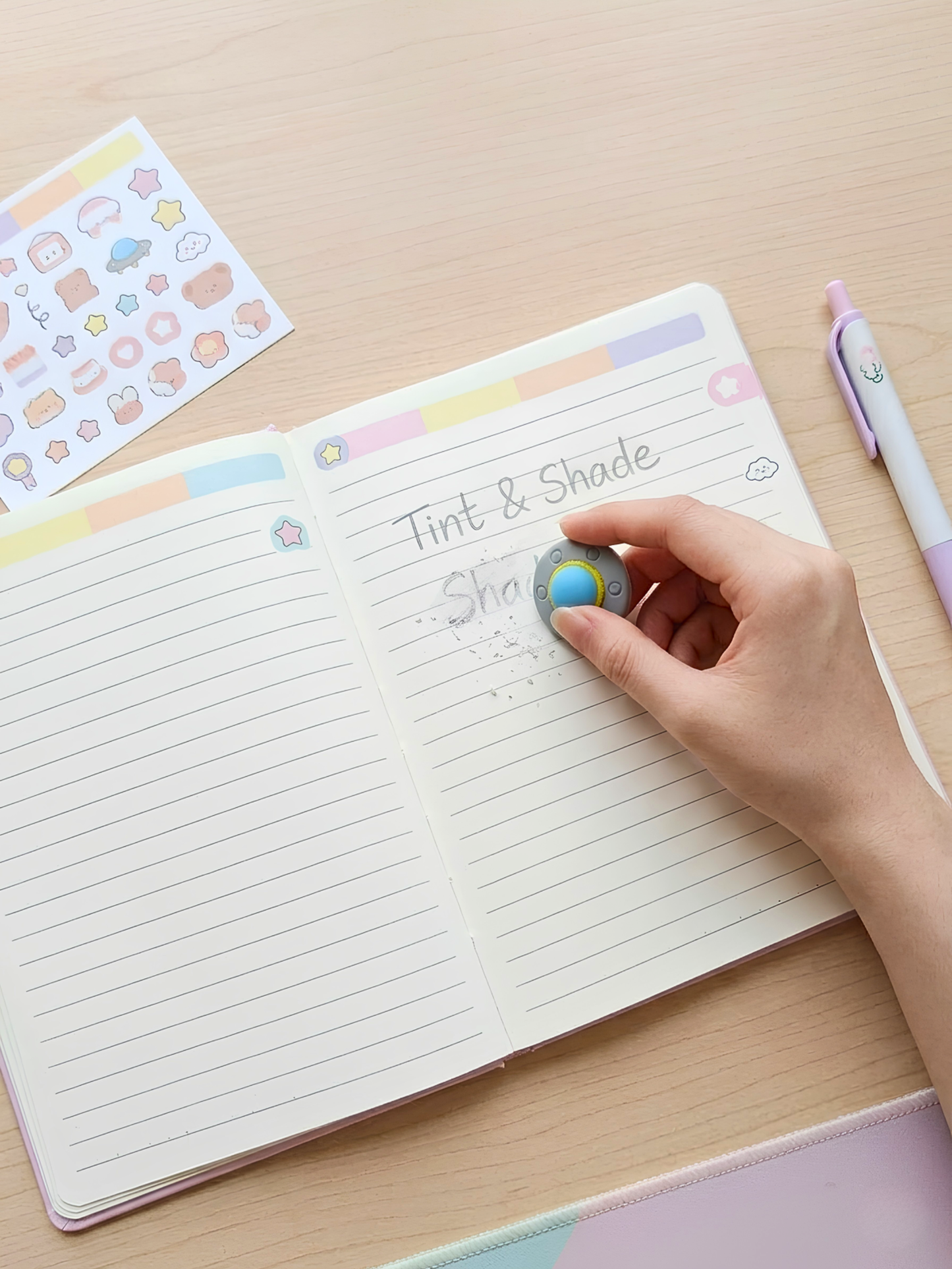Open notebook with lined pages, hand holding a small round object, and colorful stickers on a wooden surface.