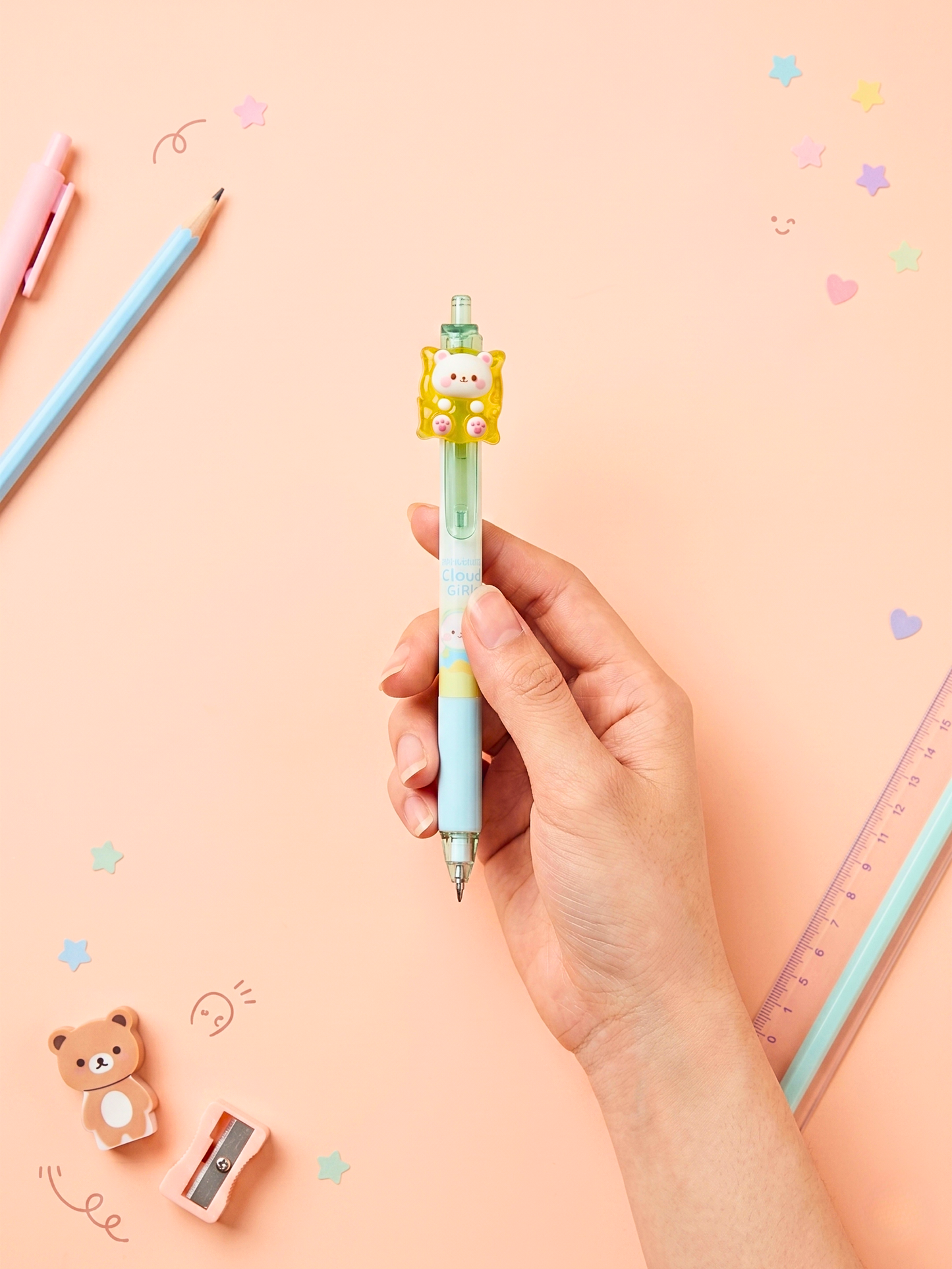 Hand holding a colorful pen with a cartoon character on a pink background with stationery items.