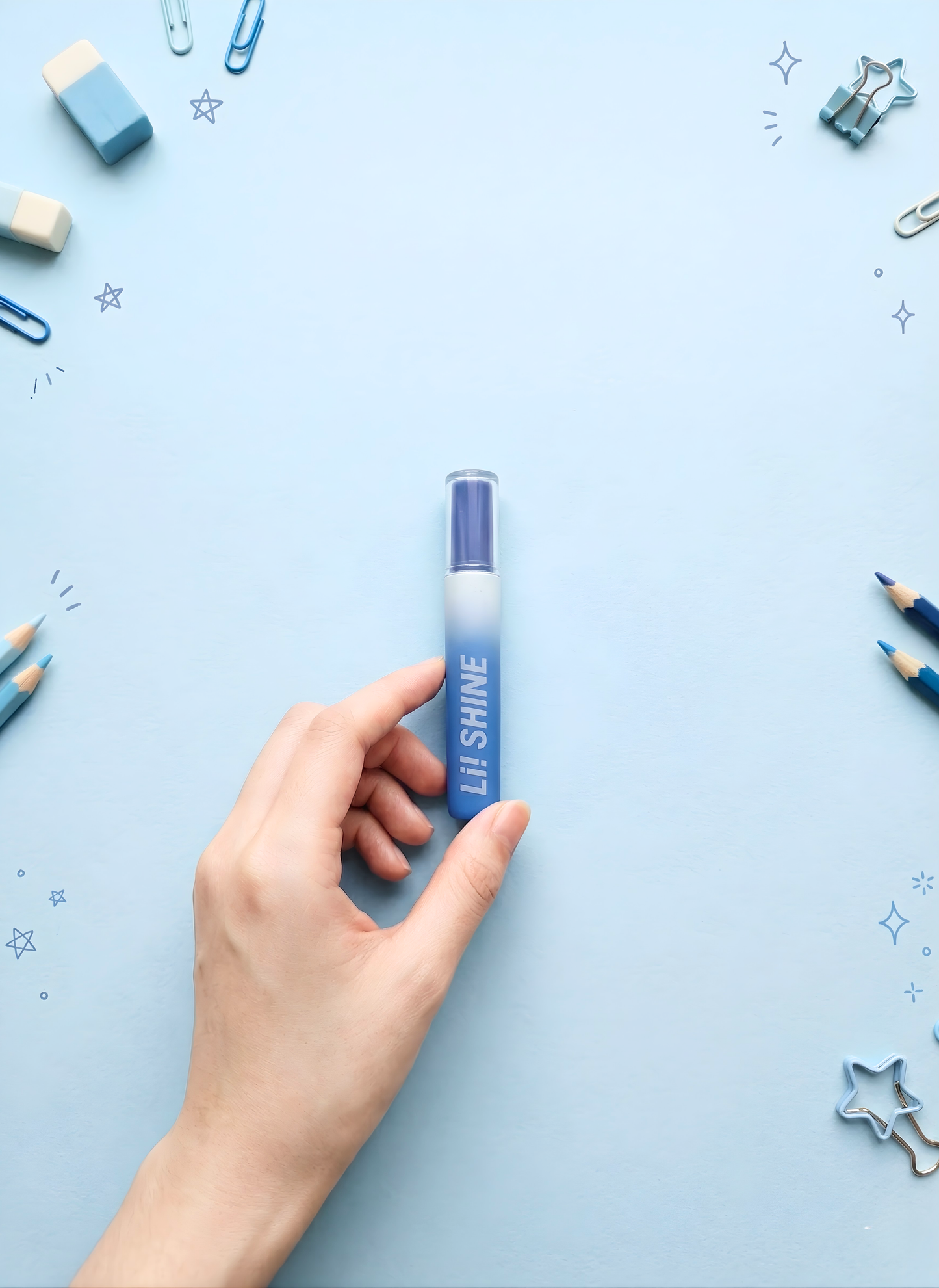 Hand holding a blue lip gloss with 'Lip Shine' text on a light blue background with stationery items.