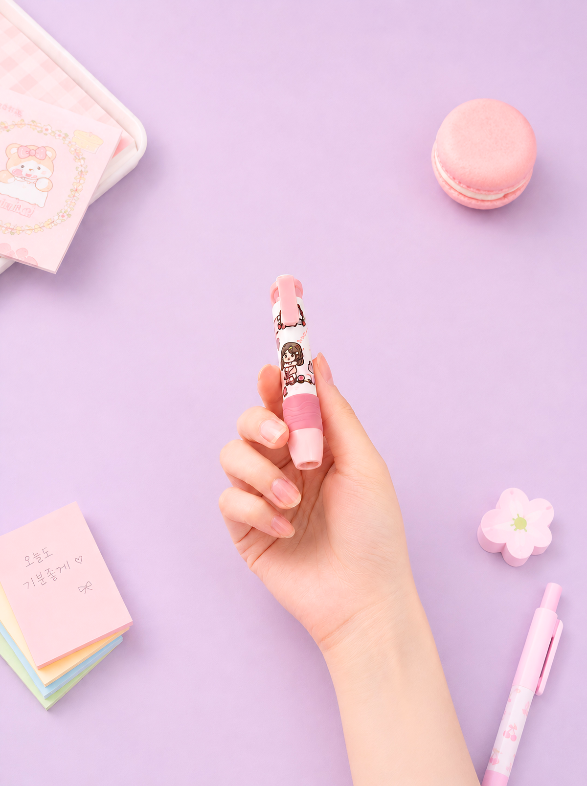 Hand holding a pink pen with a character design on a purple background with stationery items.