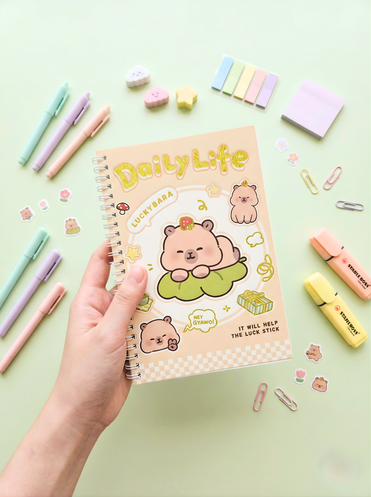 Notebook titled 'Daily Life' with cute animal illustrations held by a hand, surrounded by stationery items on a light green background.