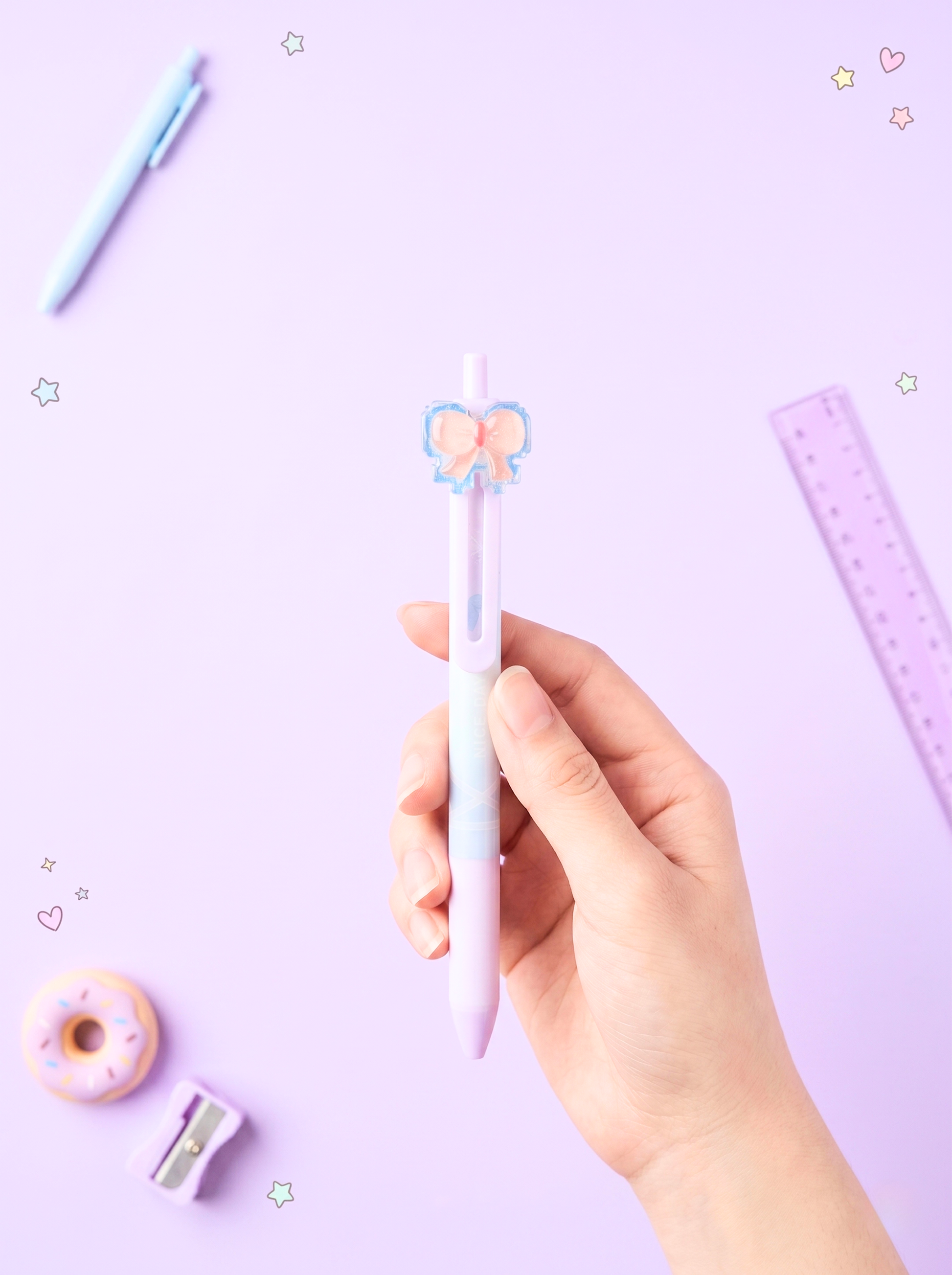Hand holding a pink pen with a butterfly design on a light purple background with stationery items.