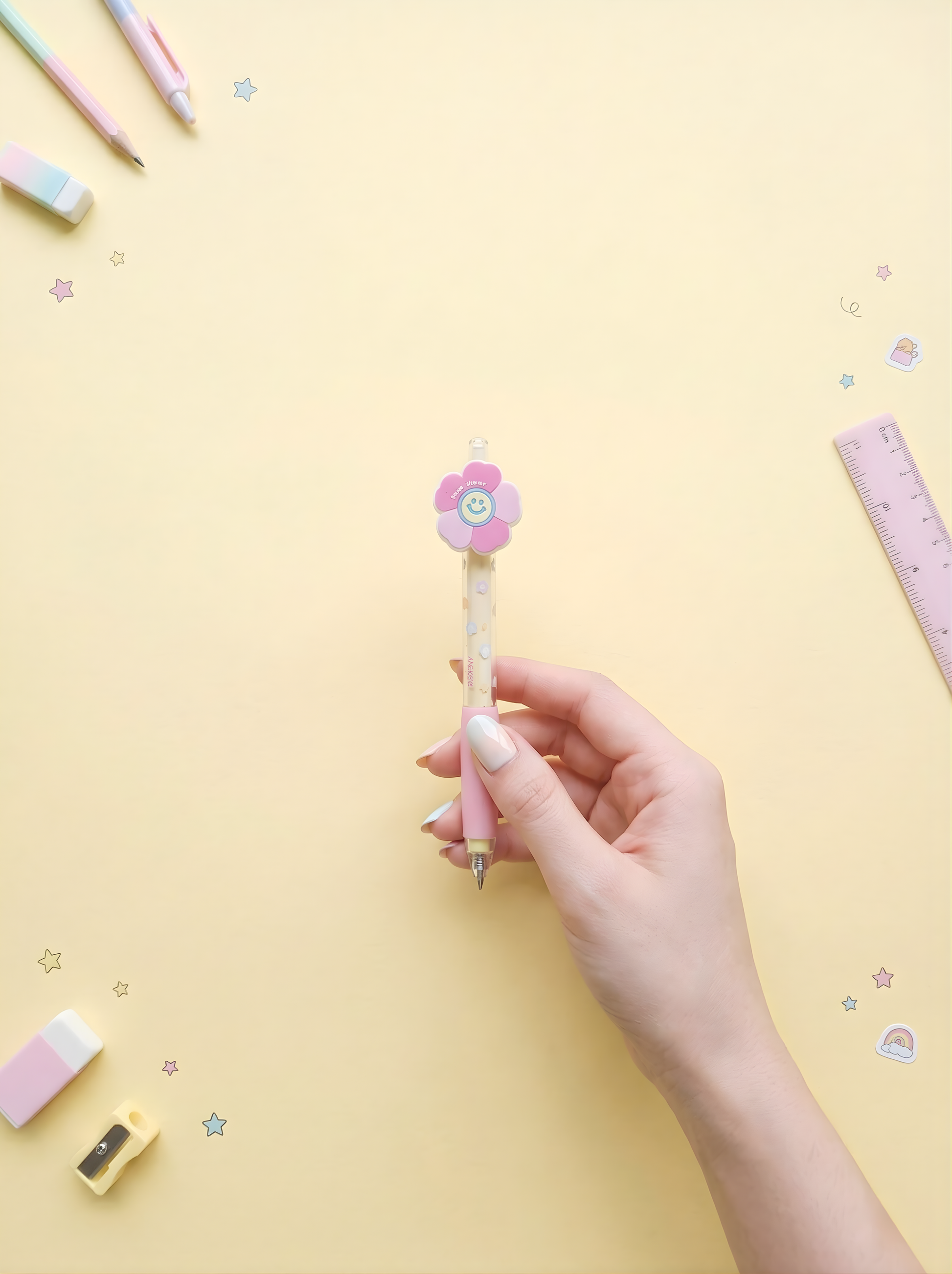 Hand holding a pink flower-shaped pen on a yellow background with stationery items.