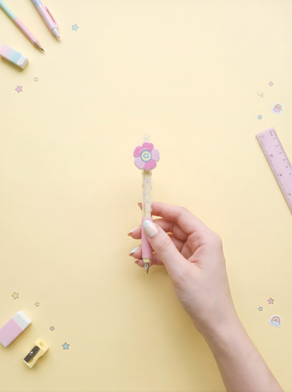 Hand holding a pink flower-shaped pen on a yellow background with stationery items.