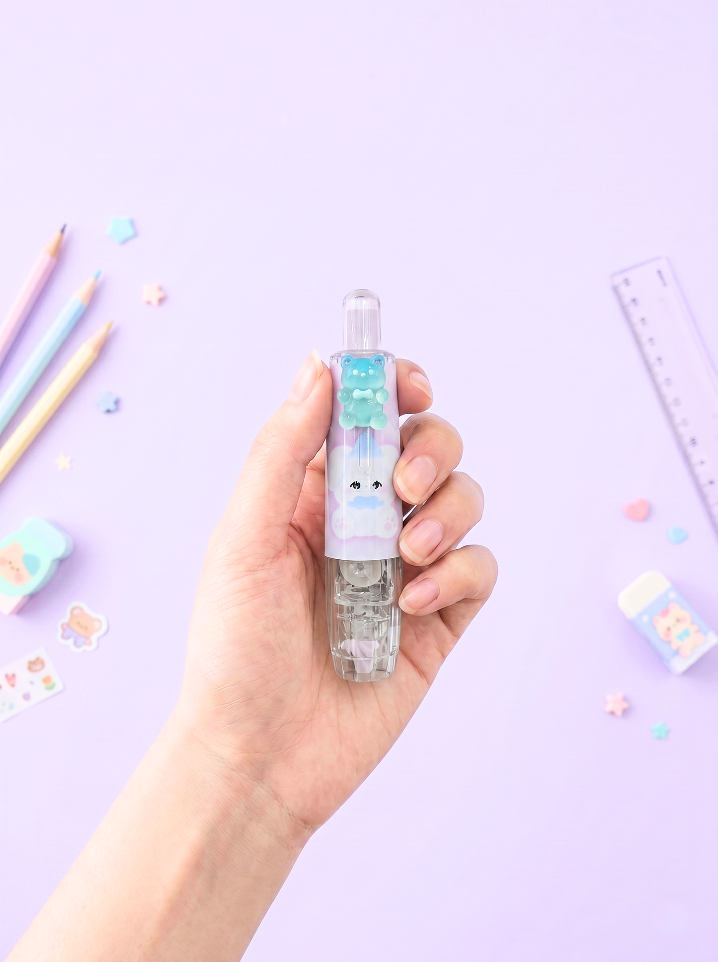 Hand holding a small bottle with a colorful cap on a light purple background with stationery items.