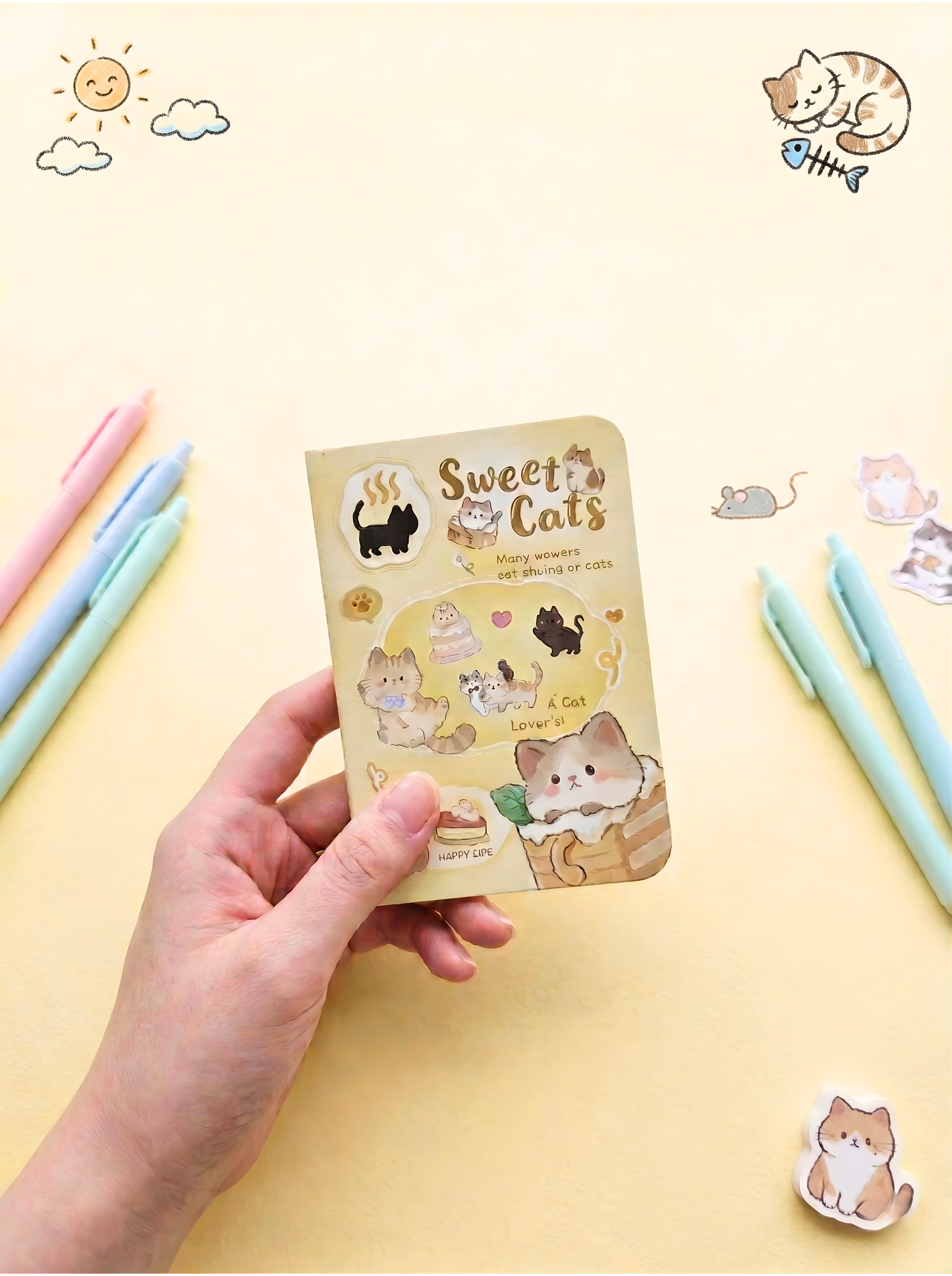 Hand holding a small notebook with cat-themed stickers on a yellow background with stationery items.