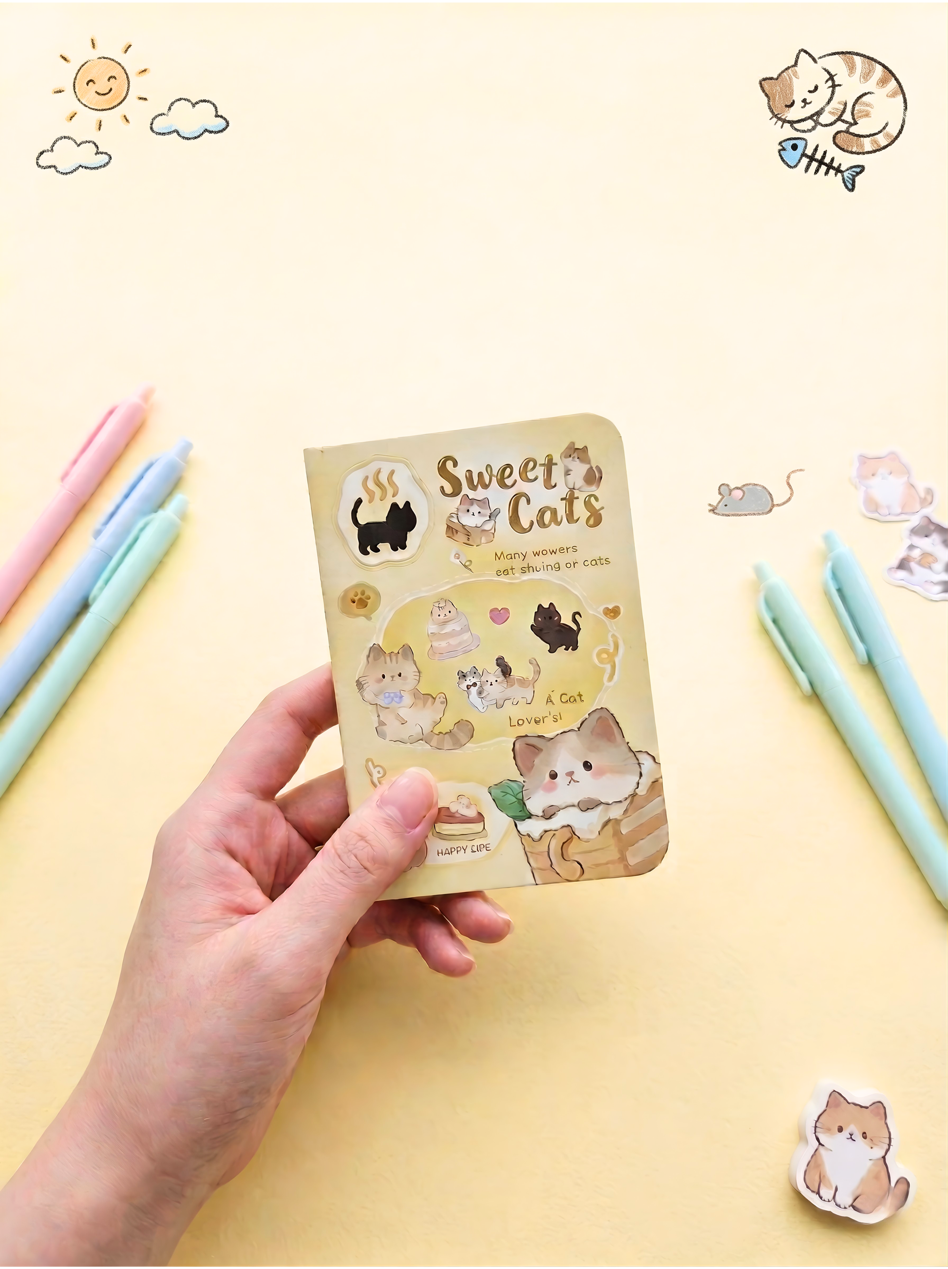 Hand holding a small notebook with cat-themed stickers on a yellow background with stationery items.